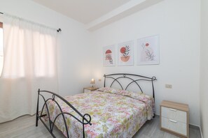 A bedroom with a bed, a nightstand, a bedside table, and wall art. - Holiday Home 'Casa Vacanze Olbia' with Shared Pool, Balcony and Air Conditioning (Le Vecchie Saline)