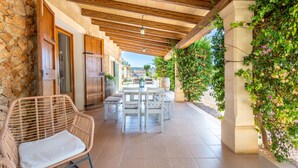 A covered patio with stone walls, wooden ceiling beams, a table with chairs, and a wicker chair with a cushion. - Country House 'Sa Vela' with Private Pool, Wi-Fi and Air Conditioning (Illes Balears)