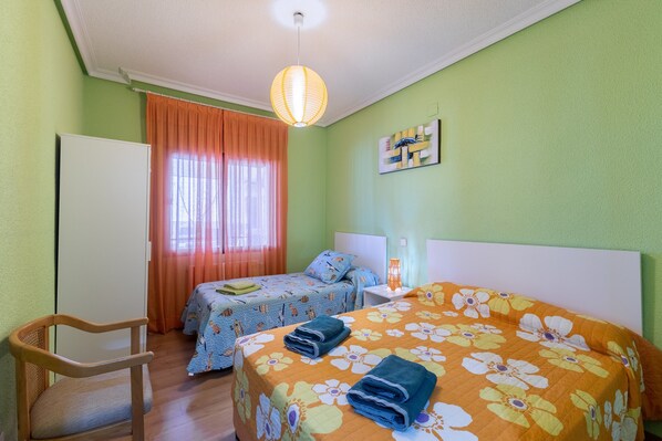 A room with two beds, a chair, a table, and a hanging light fixture.