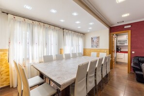 Uma sala de jantar com uma mesa comprida e cadeiras, uma cozinha ao fundo, e uma parede vermelha de destaque.