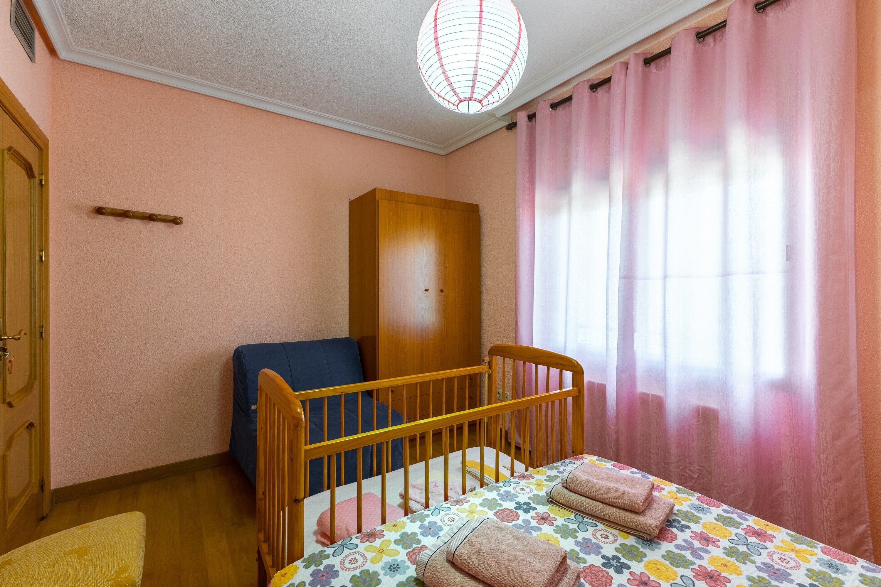 Ein Schlafzimmer mit einem hölzernen Babybett, einem Bett mit floralem Überwurf, einem Kleiderschrank und einem Fenster mit rosa Vorhängen.