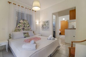 A bedroom with a bed, a bedside table, a wall-mounted light, a painting, and a kitchenette area.