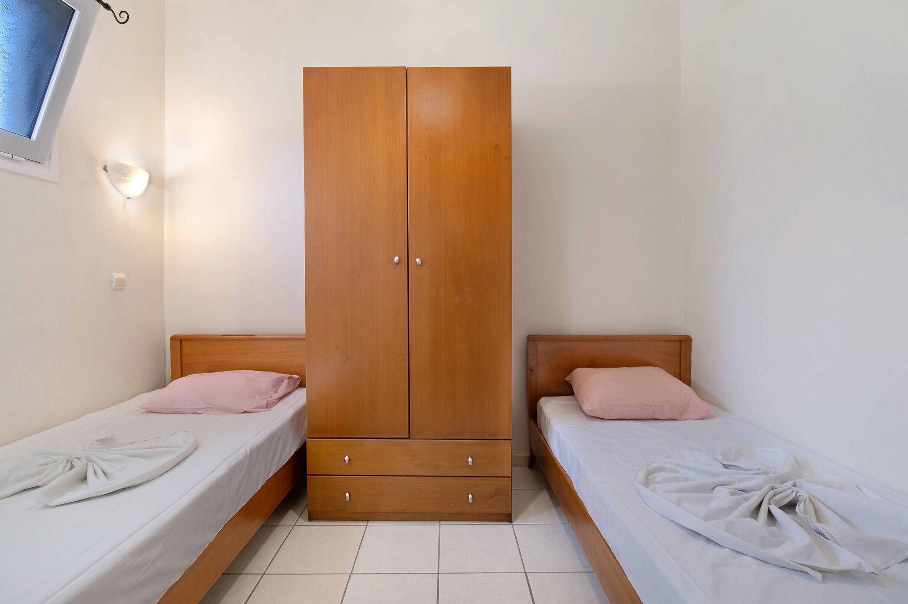 A room with two single beds, a wooden wardrobe, a bedside table, and a wall-mounted light.