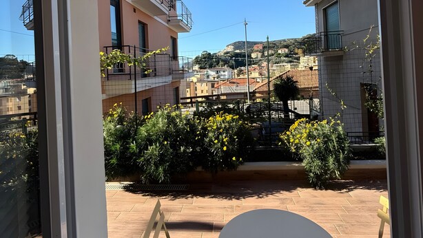 Un balcón con vista a edificios vecinos, cielo despejado y algo de vegetación.