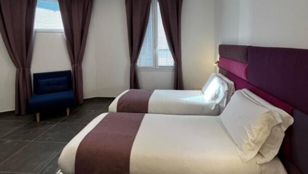 Camera d'albergo moderna con due letti, una poltrona blu e un'ampia finestra con tende.