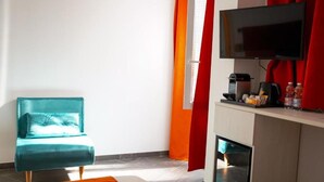 Ein modernes Hotelzimmer mit einem petrolfarbenen Sofa, einem Flachbildfernseher, einer Kaffeemaschine und einer kleinen Küchenzeile.