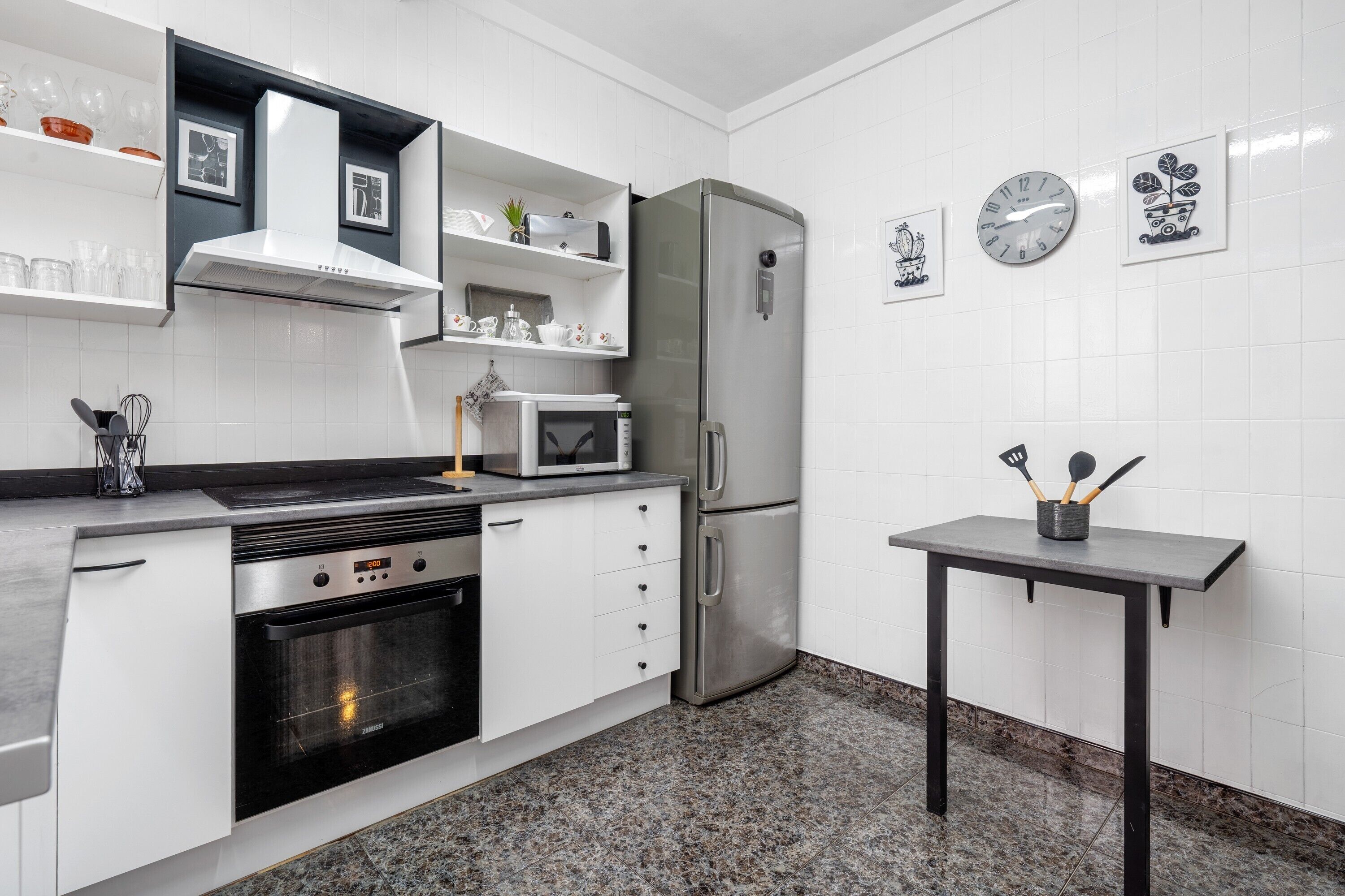 Una cocina moderna con horno empotrado, microondas, refrigerador de acero inoxidable y una pequeña mesa con utensilios de cocina.