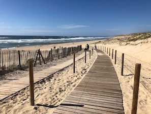 Ein hölzerner Steg führt zu einem Sandstrand, im Hintergrund sind Meereswellen zu sehen.