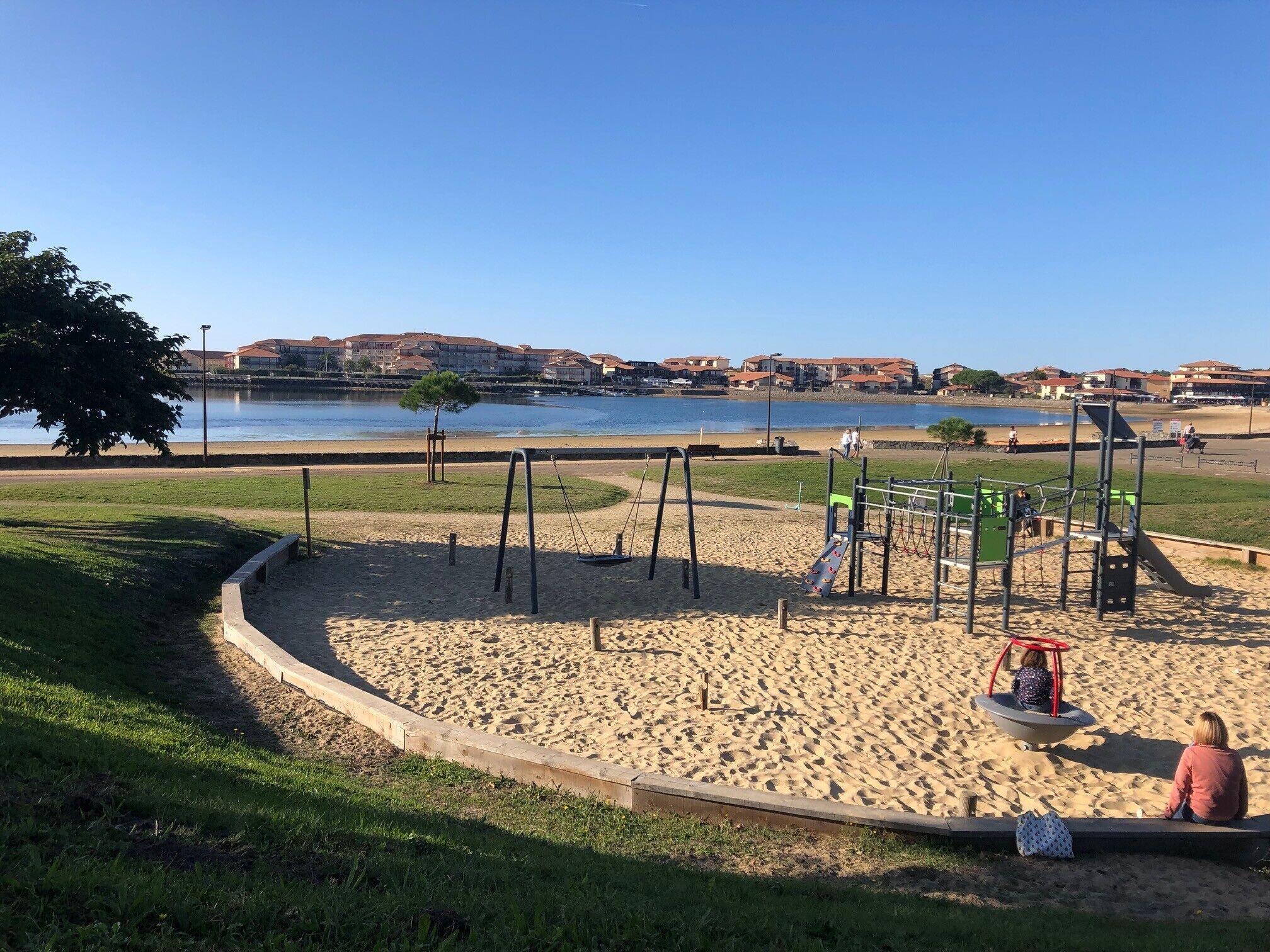 Un'area giochi con altalene e scivoli, una zona sabbiosa e un corpo d'acqua sullo sfondo.