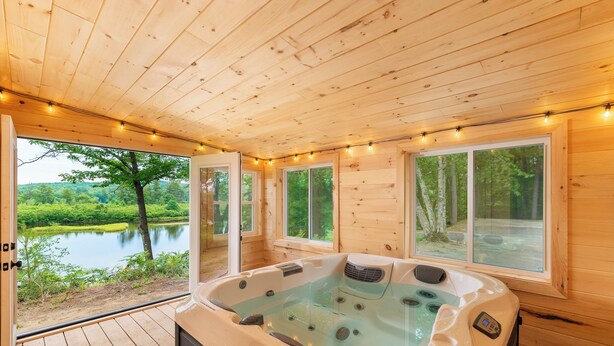 A wooden cabin with a hot tub, large windows, and a view of a lake.