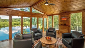 A wooden cabin with large windows overlooking a lake, furnished with leather chairs and a wooden table.