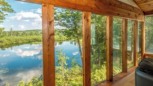 A wooden deck with large windows overlooking a calm lake surrounded by greenery.