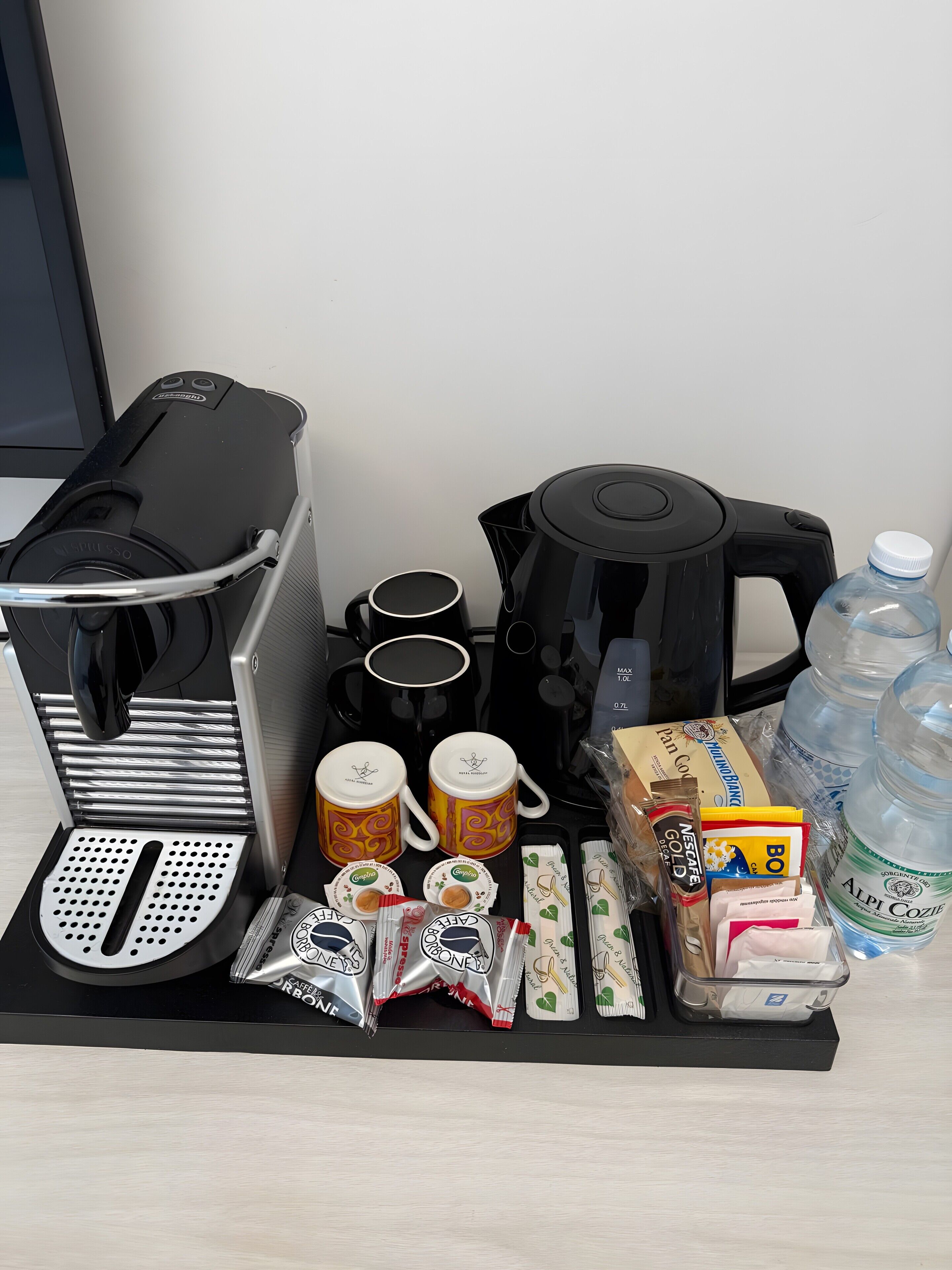 A coffee maker, two mugs, a kettle, coffee pods, a water bottle, and a box of Nescafé Gold instant coffee.