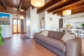 Een woonkamer met een bank, een eethoek met een tafel en stoelen, een keuken met een spoelbak en kastjes, en een hal met een deur naar buiten.
