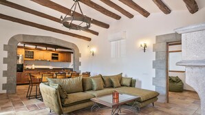 A cozy living room with a sectional sofa, a glass coffee table, exposed wooden beams, a stone fireplace, and a kitchen in the background.