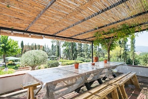 Une terrasse extérieure couverte, dotée d’une table et de bancs, et entourée de verdure et d’arbres.