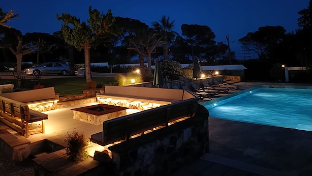 A nighttime outdoor pool area with lounge chairs, a fire pit, and a stone wall.