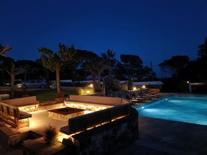 Un espace extérieur avec piscine, aménagé pour la nuit, comprenant des chaises longues, un foyer et un mur en pierre.
