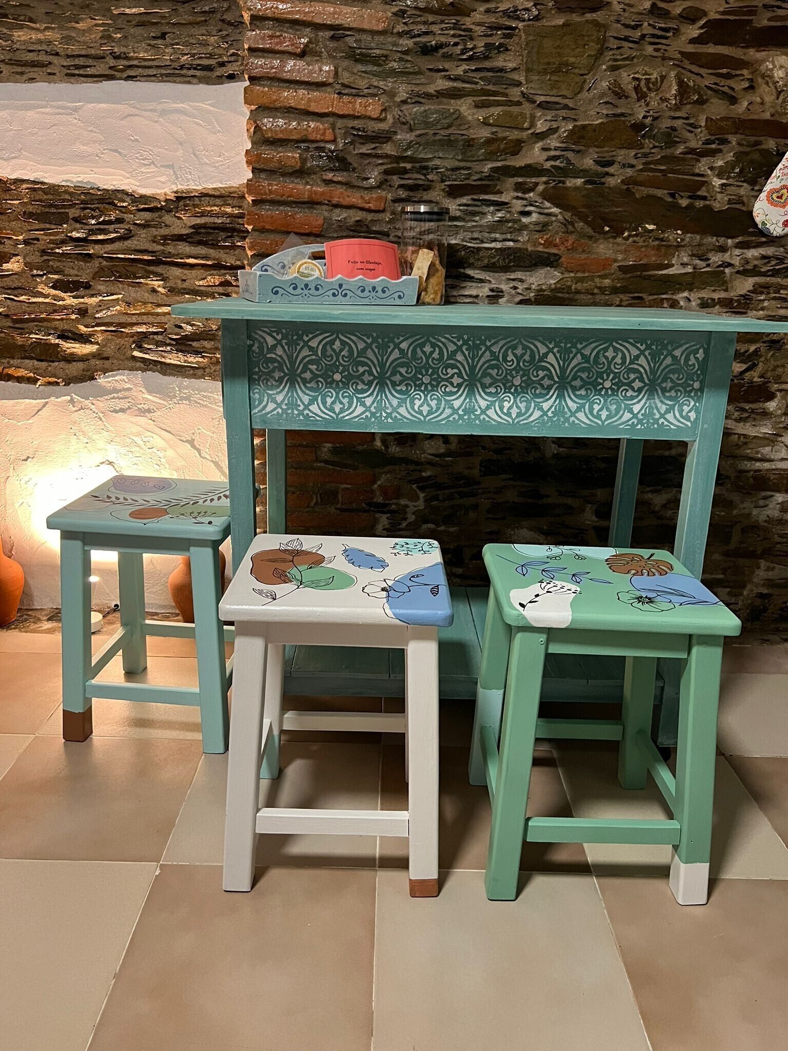 Un juego de tres sillas infantiles con estampados florales, una mesita y una pared decorativa con diseño de piedra.