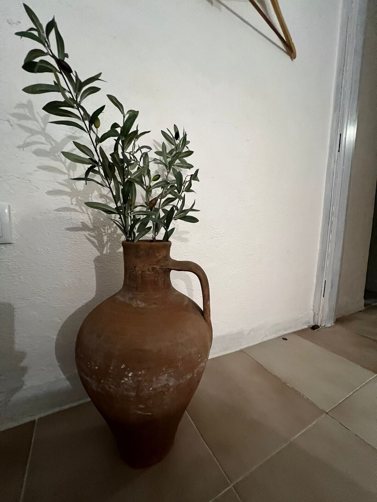 Una maceta de terracota con asa, que contiene follaje verde, sobre un suelo de baldosas frente a una pared blanca.