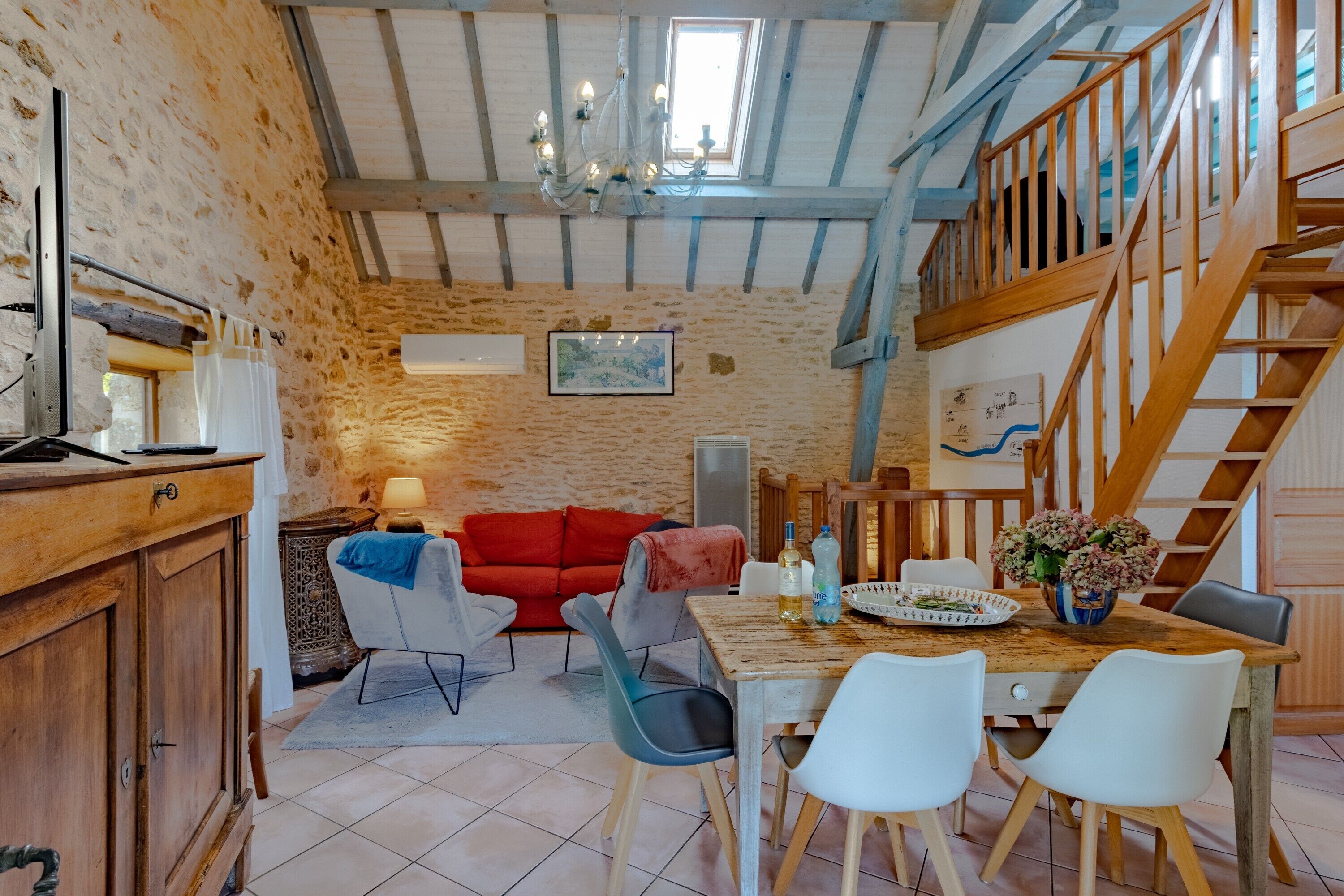 A rustic interior with exposed wooden beams, a dining area with a wooden table and chairs, a red sofa, and a staircase.
