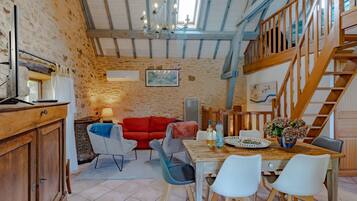 A rustic interior with exposed wooden beams, a dining area with a wooden table and chairs, a red sofa, and a staircase.