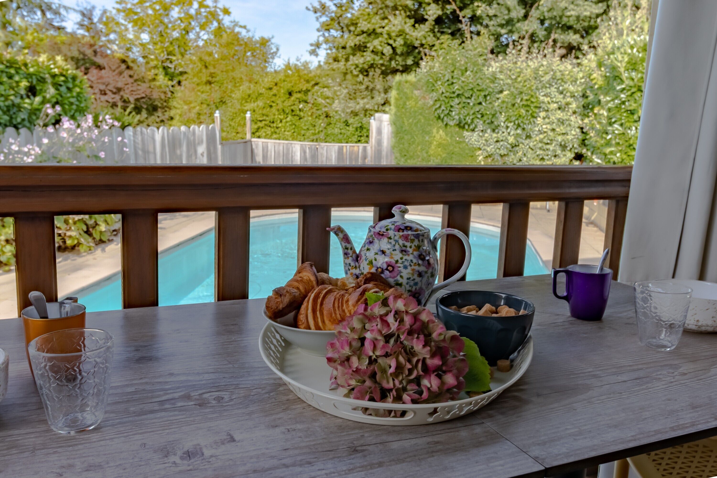 A table with croissants, a teapot, and a bowl of nuts on a balcony overlooking a pool.