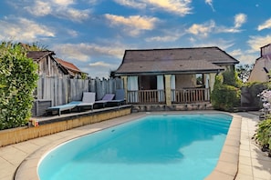 A swimming pool with a wooden deck, lounge chairs, and a small house with a balcony.