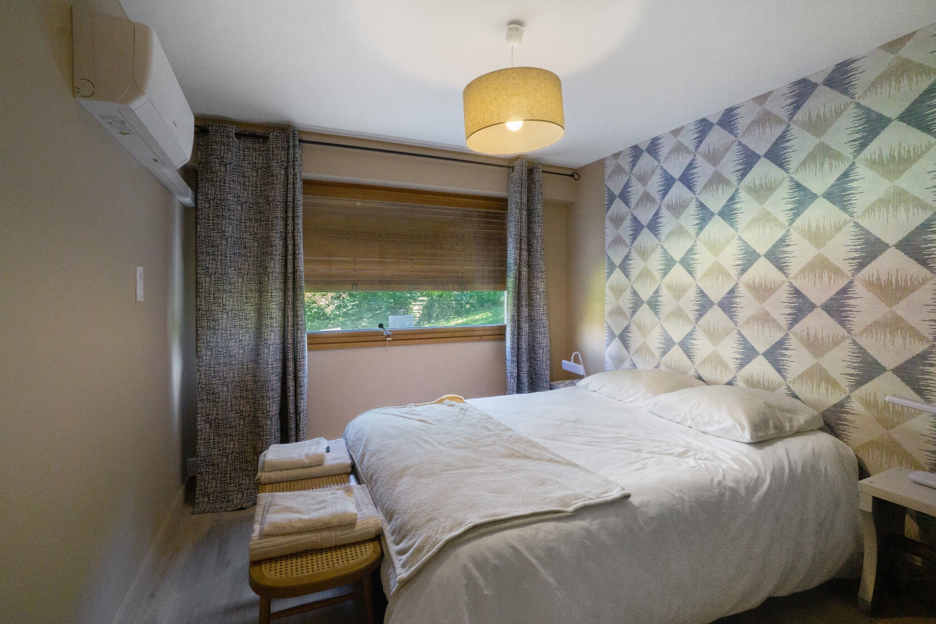 Quarto com cama, mesa de cabeceira, luminária, janela com cortinas e parede com padrão geométrico.