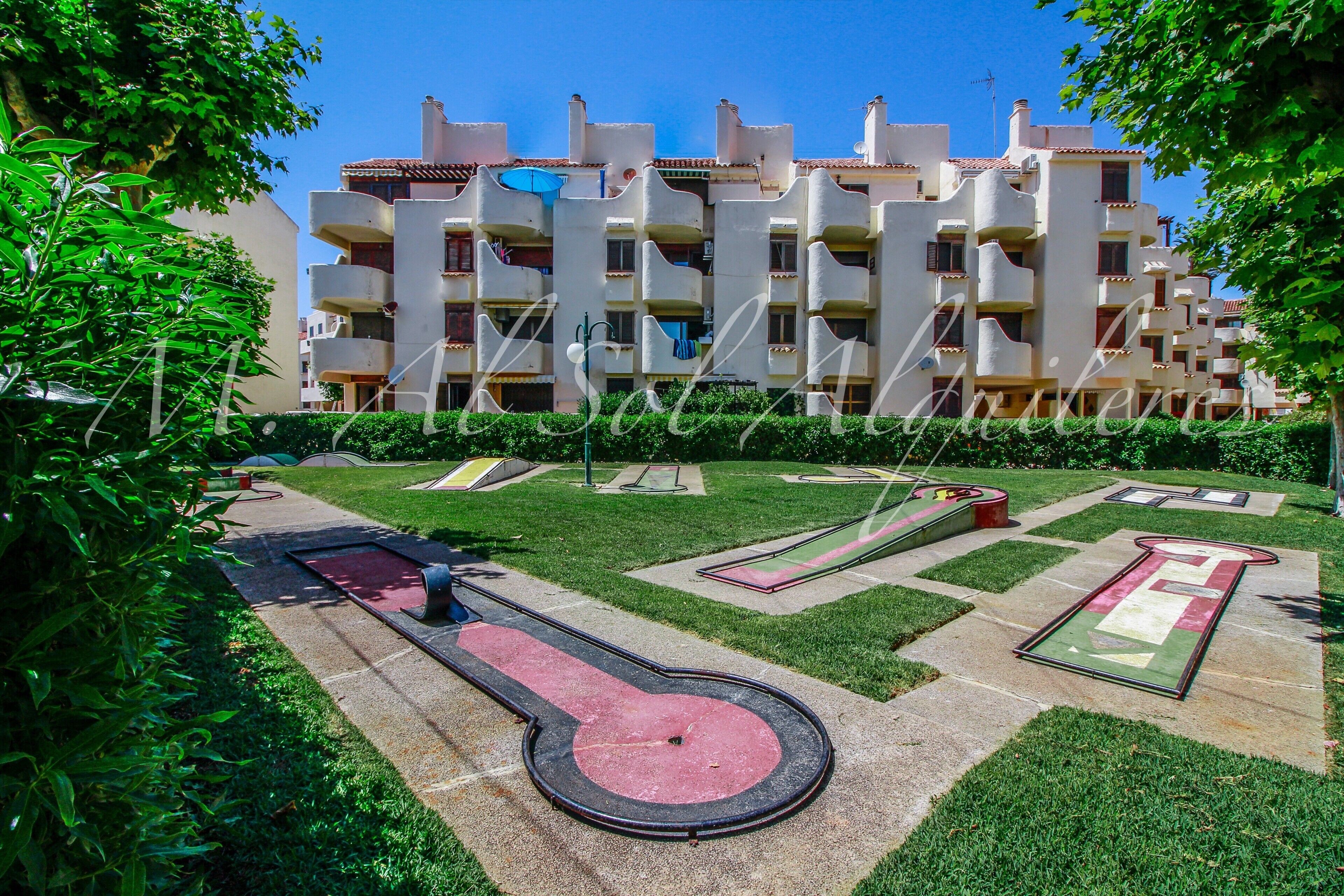 Un immeuble résidentiel doté de nombreux balcons, d’un jardin bien entretenu avec un espace de jeux pour enfants et d’un ciel d’un bleu éclatant.