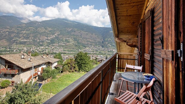 Een houten balkon met een tafel en stoelen, met uitzicht op een bergachtig landschap en een dorpje beneden.