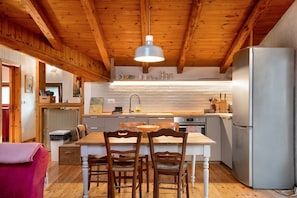 A kitchen with wooden beams, a central island with chairs, a refrigerator, and a hanging light fixture.