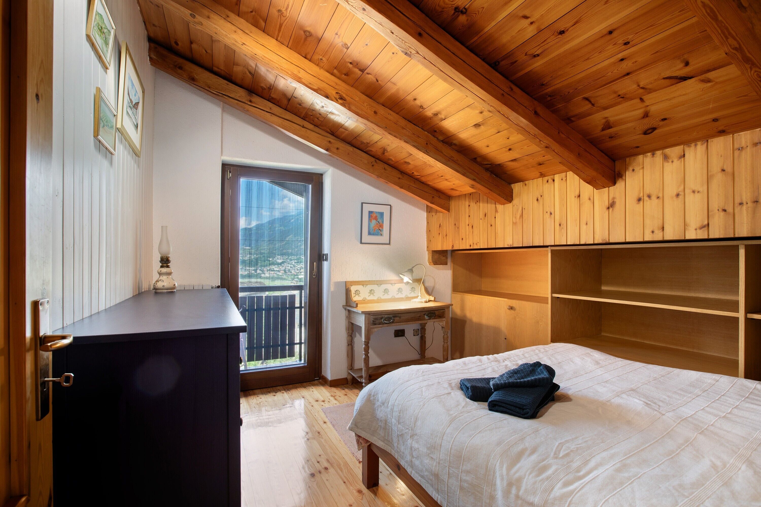 Una camera da letto con soffitto in legno, un letto con copripiumino bianco, un tavolino con una lampada e una finestra vista esterna.
