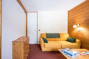 A small room with a wooden dresser, a yellow sofa with a blanket, a table with a board game, and a wooden wall with a lamp. - Apartment 'Valmorel' with Mountain View and Balcony (Les Avanchers-Valmorel)
