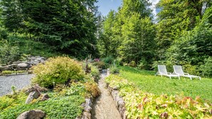 A garden with a stream, rocks, and greenery. There are two white lounge chairs in the background. - Chalet 'La Glière De L'Etelley' with Mountain View, Private Terrace and Wi-Fi (Samoëns)