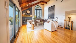 A spacious living room with wooden flooring, a fireplace, and a large window. - The View at Stoney Acres |  Pool + Hot Tub + Views (Mifflinburg)