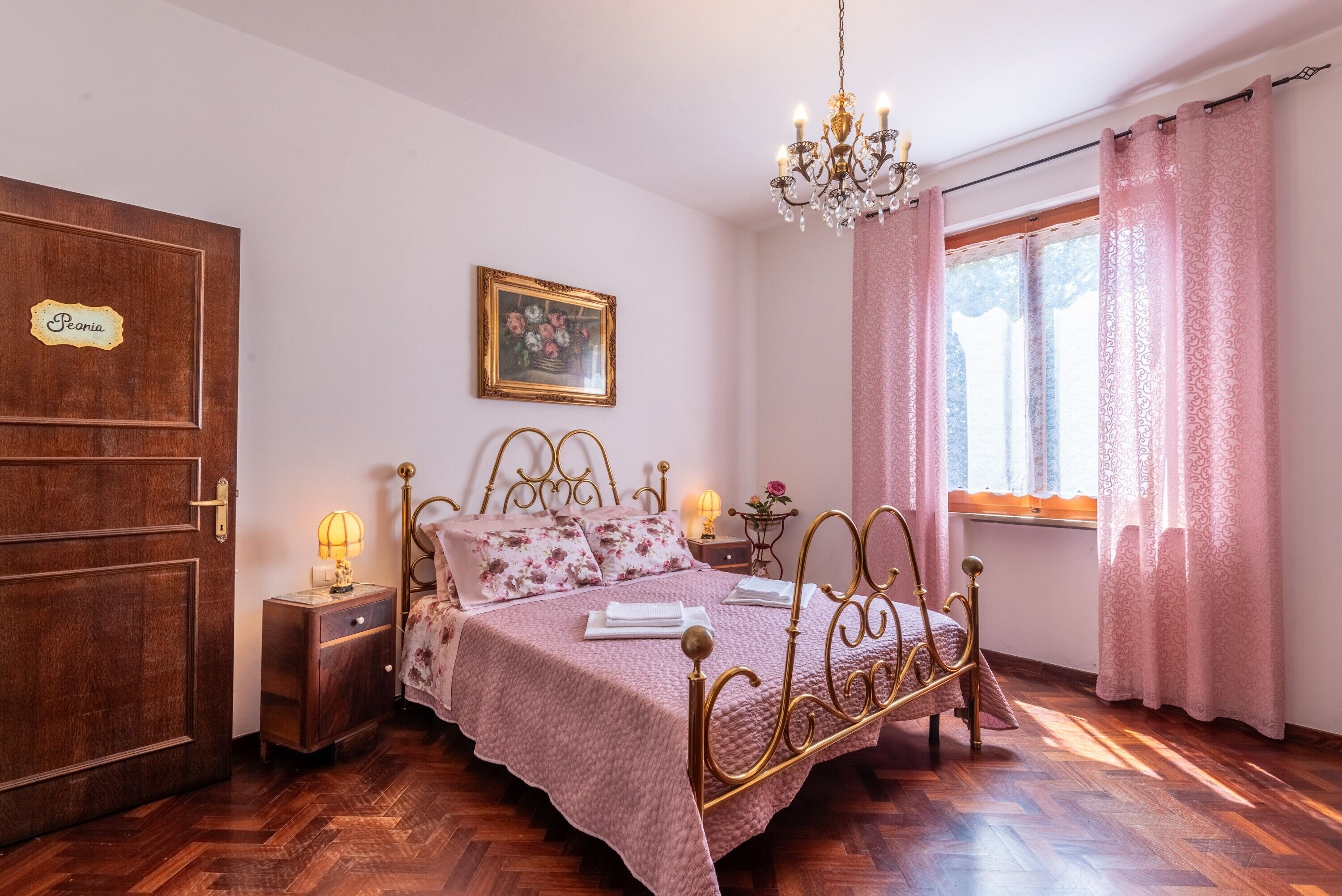 Ein Schlafzimmer mit Bett, Nachttisch, einer Kristallleuchtre, einem Fenster mit Vorhängen und einer Tür mit Namensschild.