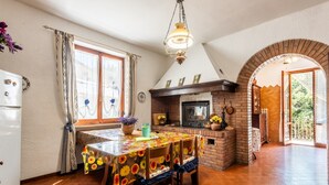 Ein Essbereich mit Tisch, der mit einer floralen Tischdecke bedeckt ist, Holzsesseln, einem Kamin aus Ziegelsteinen und einer Hängeleuchte.