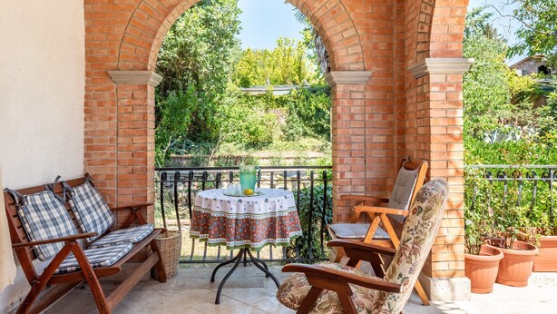 Eine Terrasse im Freien mit einem Ziegelbogen, einem runden Tisch mit Tischdecke, zwei Holzstühlen und Topfpflanzen.