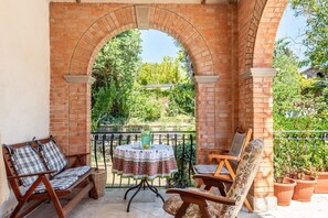 Une terrasse extérieure agrémentée d’une arche en briques, d’une table ronde recouverte d’une nappe, de deux chaises en bois et de plantes en pot.
