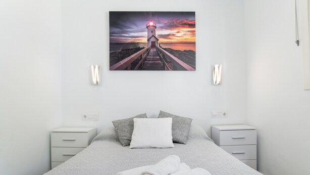 A bedroom with a bed, pillows, a folded towel, bedside tables, and a wall-mounted picture of a lighthouse at sunset.