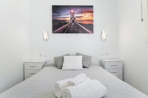 A bedroom with a bed, pillows, a folded towel, bedside tables, and a wall-mounted picture of a lighthouse at sunset.