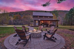 A circular fire pit with Adirondack chairs around it, a building with a porch in the background, and a hilly landscape.