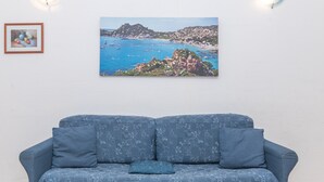 Een blauwe bank met een patroon geweven stof, een ingelijst schilderij van een kustlandschap en een klein, ingelijst foto op de muur.