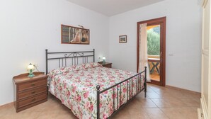 Een slaapkamer met een bed, nachtkastje, lamp, schilderij en een deur die leidt naar een buitenruimte.