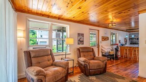 A cozy living room with two leather recliners, a wooden ceiling, and a view of the outdoors through large windows. - Frog Hollow Lodge | Riverfront, Fire Pit + Kayaks! (Jersey Shore)