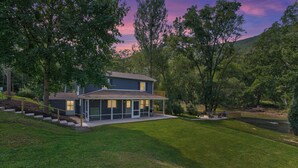 A modern two-story house with a large glass front, surrounded by lush greenery and a river. - Frog Hollow Lodge | Riverfront, Fire Pit + Kayaks! (Jersey Shore)