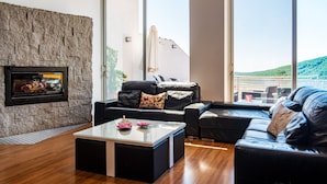 A modern living room with a stone wall, a fireplace, a black leather sofa, a glass coffee table, and a large window with a view of a balcony and greenery. - Entregredos: Rural House in Sierra de Gredos with Pool and Jacuzzi (Guisando)