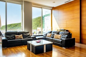 Una sala de estar moderna con un ventanal grande, un sofá de cuero negro, una mesa de centro de vidrio y piso de madera.