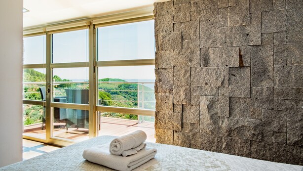 A bedroom with a large window, a stone wall, and a bed with rolled towels. - Entregredos: Rural House in Sierra de Gredos with Pool and Jacuzzi (Guisando)